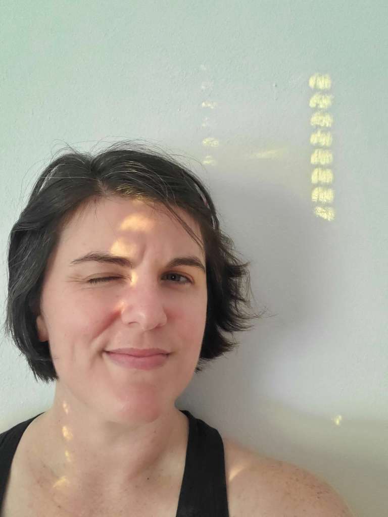 Woman with white skin and dark brown hair against a white wall. Sunlight is spotted onto woman and wall. 